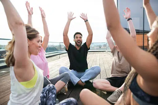 Group of people sitting in a circle on deck outdoors raising arms in a joyful activity.