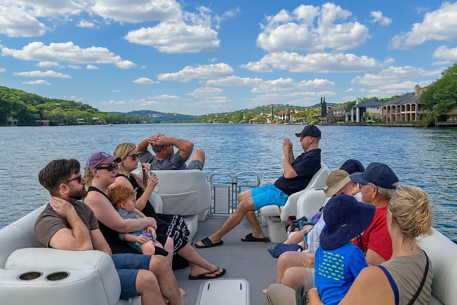 Group of families relaxing on a pontoon boat ride, enjoying scenic lake views under a blue sky with scattered clouds.