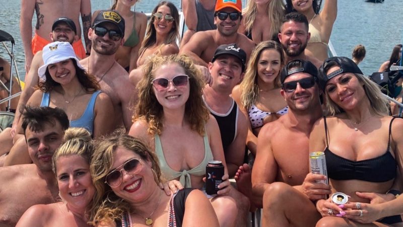 Excited bachelorette party group in matching blue swimsuits posing on a pontoon boat with drinks.