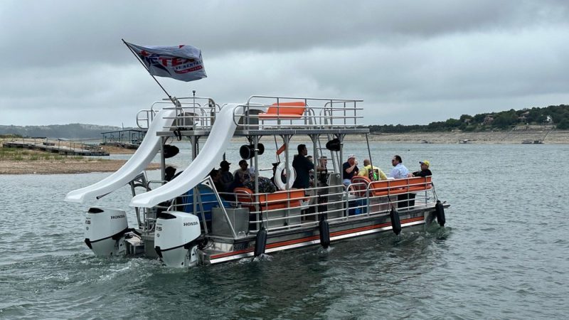 Pontoon boat with waterslide carrying a group of people enjoying a sunny day on the lake.
